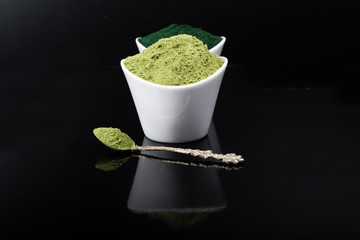 An isolated tablespoon of dried organic wheat grass and spirulina powder, on white rustic background