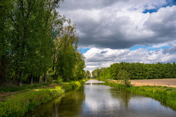 Fototapeta na wymiar Canal with overhanging clouds and trees to the left in the spring. Farm land on the lefthandsite.