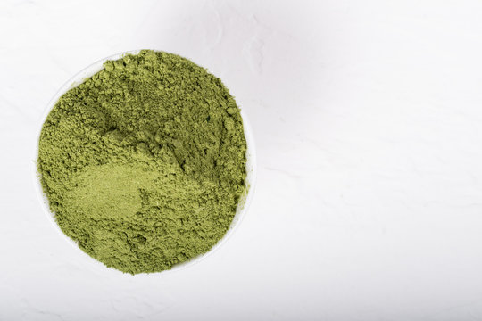 An isolated tablespoon of dried organic wheat grass and spirulina powder, on white rustic background