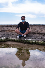 Fototapeta na wymiar man on the beach sit on rock near a sea wearing mask for protect against infection with virus or coronavirus, civid19. enjoy sea in 2020 concept. person alone in black shirt with reflection in water
