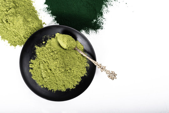 An isolated tablespoon of dried organic wheat grass and spirulina powder, on white rustic background