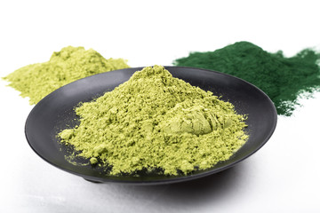 An isolated tablespoon of dried organic wheat grass and spirulina powder, on white rustic background