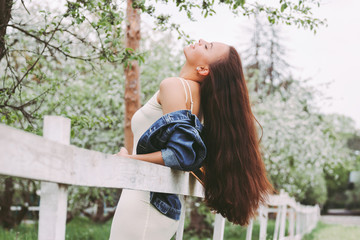 Naklejka premium Portrait young carefree hippie girl in denim jacket with long brunette hair on countryside garden among blossom trees. Beautiful happy hipster woman in stylish dress posing with hair on green meadow