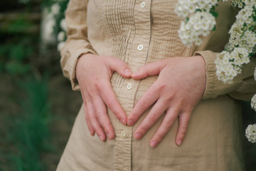 pregnant woman outdoors and makes a heart-shaped hands on her belly the background of flowers 