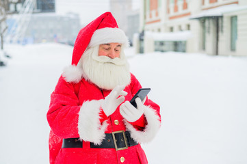 Surprised santa claus is using mobile outside. Santa Claus stands in the background of buildings in winter and calls on a smartphone. Snow in December.
