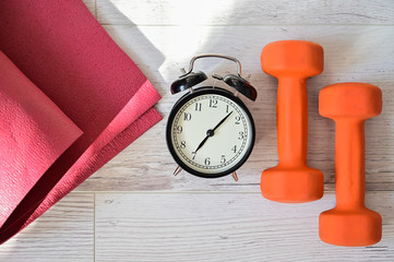 Top view on orange dumbbell alarm clock and raspberry yoga mat. The concept of home sports during quarantine. Fitness time. Early rise for classes.