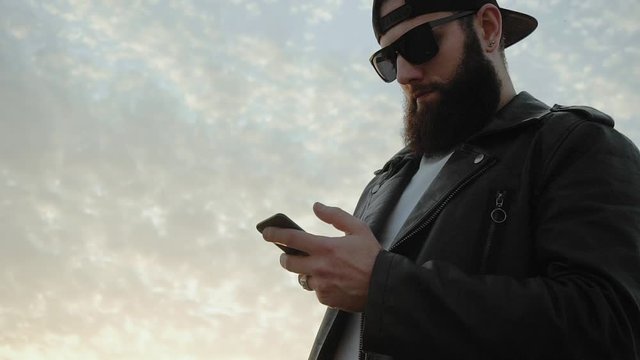 Bearded brutal image guy looks at smartphone and watching news feed. Man places order in online store and makes purchase payment. Communication. Stylish man moves finger across screen. Remote Shoping
