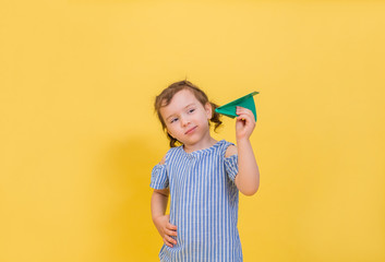 Cute blonde girl on a yellow background with a green origami airplane. A little girl in a striped t-shirt on a yellow background with a copy of space. military air force day.