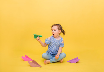 Cute blonde girl launches an origami airplane on a yellow background with a copy of space. A little girl is sitting with colorful airplanes. learning origami.