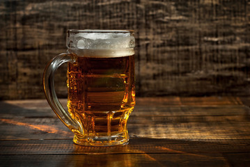 A glass of amber beer. Glasses on a wooden table. Black shabby boards on the background. Bright contrasting light and glare on the glass. Place for text near the booze.