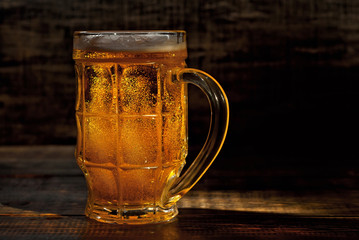 A glass of amber beer. Glasses on a wooden table. Black shabby boards on the background. Bright contrasting light and glare on the glass. Place for text near the booze.