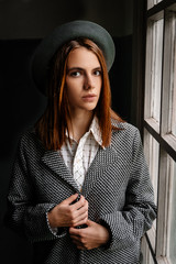 Beautiful young  pensive girl with long hair, wears a stylish hat, a gray coat, a white shirt and pants, poses for an outdoor portrait in daylight, showing a female spring and autumn fashion concept.