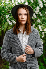 Beautiful young  pensive girl with long hair, wears a stylish hat, a gray coat, a white shirt and pants, poses for an outdoor portrait in daylight, showing a female spring and autumn fashion concept.