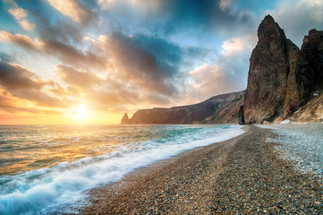 Bright sea sunset. The waves crash into the rock, lit by the warm sunset, sand and pebbles,...