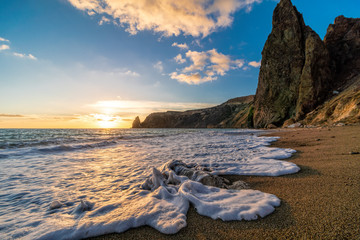 Bright sea sunset. The waves crash into the rock, lit by the warm sunset, sand and pebbles,...