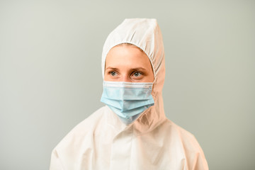 Portrait of young medic woman in white protective suit with hood on gray background.
