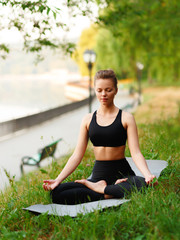 Young attractive woman practicing yoga, sitting in Ardha Padmasana exercise, Half Lotus pose, working out, wearing in black sportswear meditation session at the park. Full length.