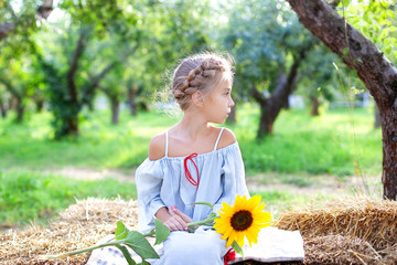 Little girl with pigtail on her head sits on roll of haystacks in garden and holds sunflower. Child sits on straw and enjoys nature in countryside. Childhood concept. Child is sad. Loneliness. Pensive