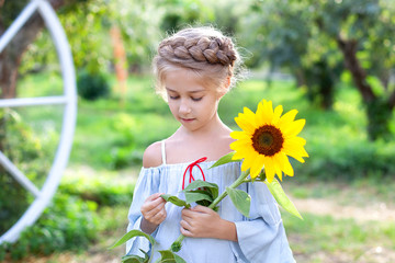 Smiling little girl with a pigtail on her head holds sunflower in garden. Childhood. Child with sunflower. Child enjoying nature on sunny summer day. Closeup portrait young blonde girl with sunflower.