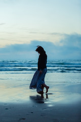 A sad woman with long hair in a long dress is walking at sunset along the shore of the sea or ocean. Social exclusion