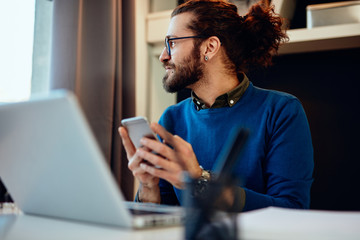 Young attractive Caucasian hipster sitting in his office and using smart phone.