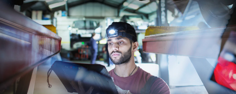 Focused male mechanic with headlight and diagnostic equipment working under car in auto repair shop