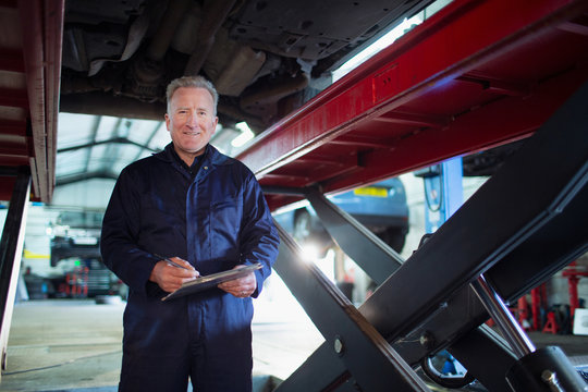 Portrait confident, smiling male mechanic with clipboard working under car in auto repair shop