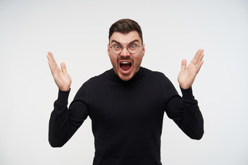 Angry young short haired unshaved brunette guy in black poloneck raising his hands and frowning face while screaming crossly, isolated over white background