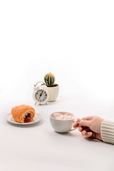 A female hand holds a cup of coffee with marshmallows, on a saucer lies a croissant with jam and there is an alarm clock with cactus.
