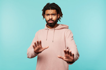 Frightened young dark skinned bearded male with dreadlocks raising hands in protective gesture and looking scaredly at camera, posing over blue background