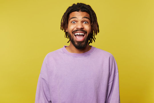 Joyful young attractive dark skinned brown haired man with dreadlocks looking surprisedly at camera with opened mouth while standing over yellow background