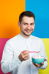 A man on a yellow, blue, orange, pink background in a white shirt with a turquoise plate of marshmallows and a pink spoon. The guy with a smile. An image for a male photo. Photo session concept