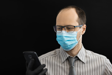 Young man in a surgical mask and a tie looks at the phone. Black background.