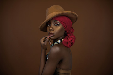 A sensual looking young black female with beautiful makeup posing by herself in a studio wearing a brown strapless top, red head scarf, brown hat and gorgeous colorful necklace.