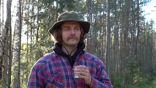 portrait of hipster millennial caucasian male with moustache in green panama hat drinks hot tea or specialty coffee in morning, mountain forrest camping 