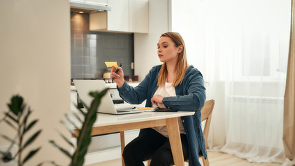 Using sticky notes. Young caucasian girl in casual clothes working from home, she is sitting at kitchen table at home and using laptop, looking at sticky note