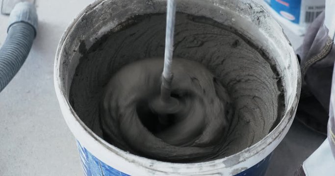 Mixing cement glue or mortar with long drill. Worker mixing cement glue in bucket, cement mixer in construction site, close up, at construction site in plastic bucket man carefully mixes mortar