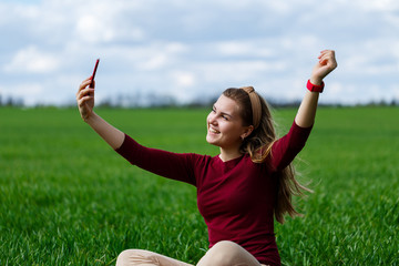 Beautiful woman with a phone in her hands sits on the grass. The girl looks at herself in the camera of the phone and takes selfie photos. She smiles and enjoys a warm day. Concept photo on smartphone