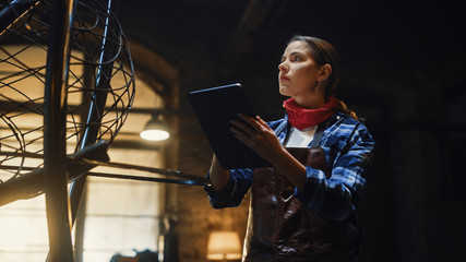 Beautiful Female Artist Sketches on a Tablet Computer Next to Brutal Metal Sculpture in Studio. Tomboy Girl Wears Checkered Shirt and Apron. Contemporary Fabricator Creating Abstract Steel Art.