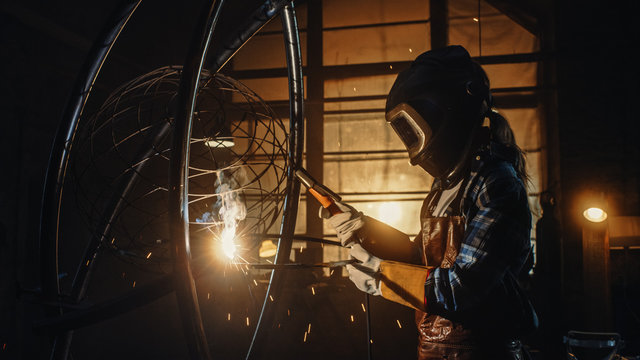 Beautiful Female Artist is Welding a Brutal Metal Sculpture in a Dark Studio. Tomboy Girl Polishes Metal Tube with Sparks Flying Off It. Contemporary Fabricator Creating Abstract Steel Art.