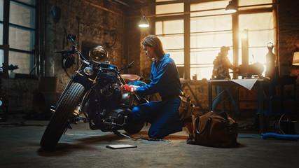 Young Beautiful Female Mechanic is Working on a Custom Bobber Motorcycle. Talented Girl Wearing a Blue Jumpsuit. She Uses a Ratchet Spanner to Tighten Nut Bolts. Creative Authentic Workshop Garage.