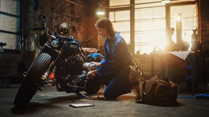 Young Beautiful Female Mechanic is Working on a Custom Bobber Motorcycle. Talented Girl Wearing a Blue Jumpsuit. She Uses a Ratchet Spanner to Tighten Nut Bolts. Creative Authentic Workshop Garage.