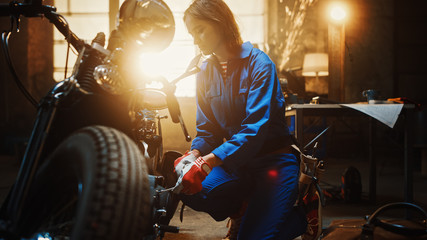 Obraz na płótnie Canvas Young Beautiful Female Mechanic is Working on a Custom Bobber Motorcycle. Talented Girl Wearing a Blue Jumpsuit. She Uses a Spanner to Tighten Nut Bolts. Creative Authentic Workshop Garage.