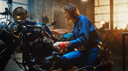 Fototapeta na wymiar Young Beautiful Female Mechanic Comes Working on a Custom Motorcycle in Garage. Talented Girl Wearing a Blue Jumpsuit. She Uses a Ratchet to Tighten Nut Bolts. Creative Authentic Workshop.