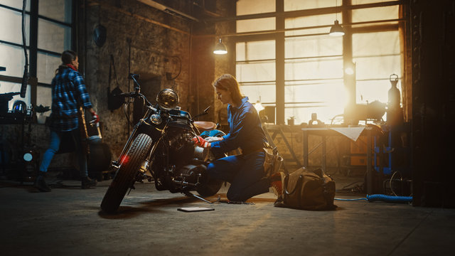 Young Beautiful Female Mechanic is Working on a Custom Bobber Motorcycle. Talented Girl Wearing a Blue Jumpsuit. She Uses a Ratchet Spanner to Tighten Nut Bolts. Creative Authentic Workshop Garage. © Gorodenkoff