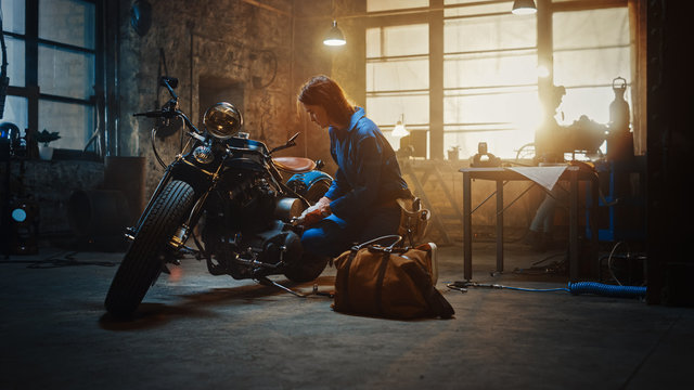 Young Beautiful Female Mechanic Comes to Garage and Starts Working on a Custom Motorcycle. Talented Girl Wearing a Blue Jumpsuit. She Uses a Spanner to Tighten Nut Bolts. Creative Authentic Workshop. © Gorodenkoff