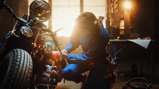 Young Beautiful Female Mechanic is Working on a Custom Bobber Motorcycle. Talented Girl Wearing a Blue Jumpsuit. She Uses a Spanner to Tighten Nut Bolts. Creative Authentic Workshop Garage.