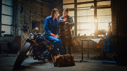 Two Young Beautiful Female are Discussing The Work Done on a Custom Bobber Motorcycle. Talented Girls Use a Tablet Computer. They are In Good Spirit and Happy. Creative Authentic Workshop Garage.