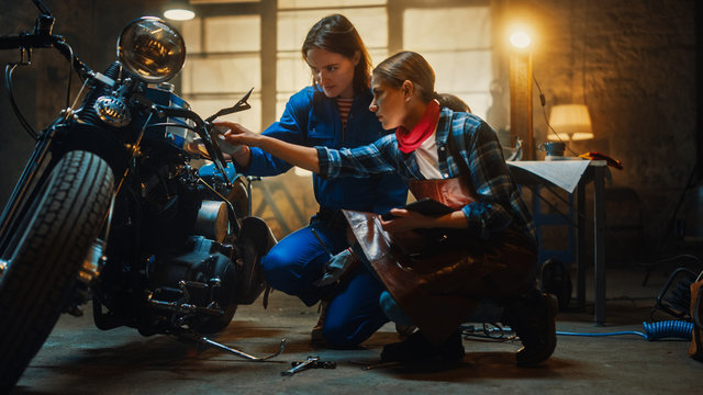 Two Young Beautiful Female are Discussing The Work Done on a Custom Bobber Motorcycle. Talented Girls Use a Tablet Computer. They are In Good Spirit and Happy. Creative Authentic Workshop Garage. - Powered by Adobe