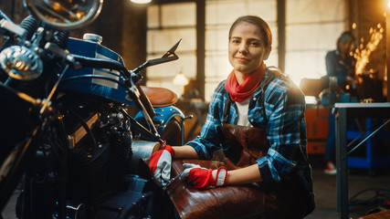Young Beautiful Female Mechanic is Working on a Custom Bobber Motorcycle. Talented Girl Wearing a Checkered Shirt and an Apron. She Smiles at the Camera. Creative Authentic Workshop Garage. © Gorodenkoff
